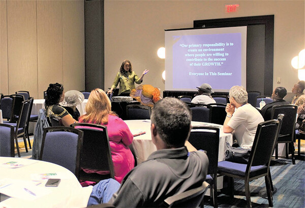 Pg9CarolynMcCallPryorLearning.jpg.jpg A woman standing in front of a screen with a PowerPoint showing while speaking to a room full of people seated at tables