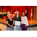 Three women posing with an award in front of the stage with various flags in the background.