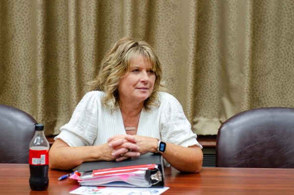 A woman with her hands linked sitting at a table listening to another individual out of frame speak.