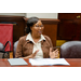 A woman with glasses sitting at a table while speaking and gesturing with her hands.