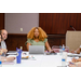 A woman at the head of the table with a laptop listening to someone speak at the other end of the table out of frame.