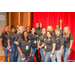 A big group photo of individuals standing in front of a red curtain and two flags.