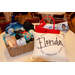 A table with various gift items on top: a pillow, a red hand bag, and a brown gift basket with candles, gift cards, and a hat.