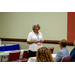 A woman standing in front of a table speaking to the individuals that are sitting down in front of her.