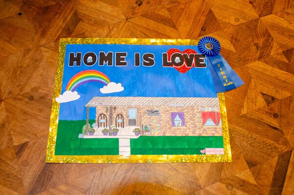 A first place What Home Means to Me poster with a brick house, a rainbow, and Home is love written at the top.