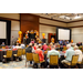 A room full of individuals sitting at tables with with several balloon arches is in the background.