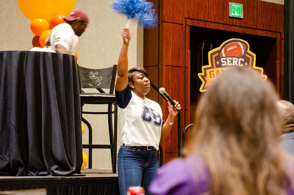 A woman in a shirt with the number 88 on it holding up a blue pom pom while speaking into a microphone.