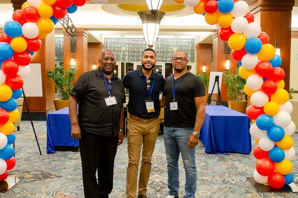 Three men taking a group photo under a balloon arch.