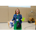 A woman in a green dress and jean jacket holding up her Bingo card in one hand and a gift bag in the other.